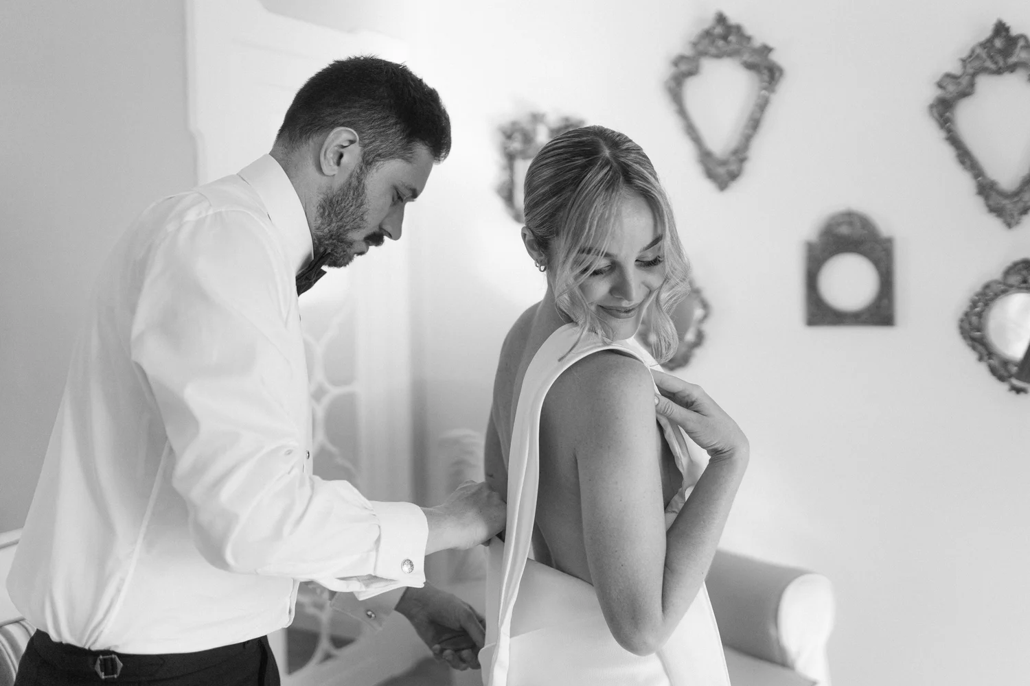 A man helping a woman try on a wedding dress in a bridal boutique, with decorative mirrors on the wall behind them.