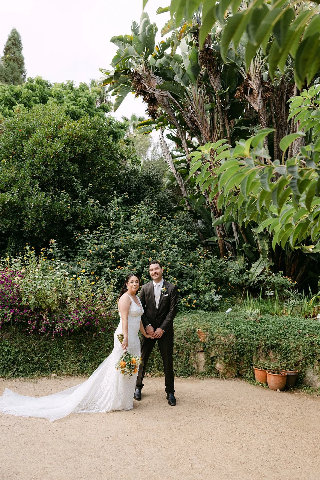 A bride and groom standing outdoors in a lush garden, smiling, holding hands, with the bride holding a bouquet of flowers.