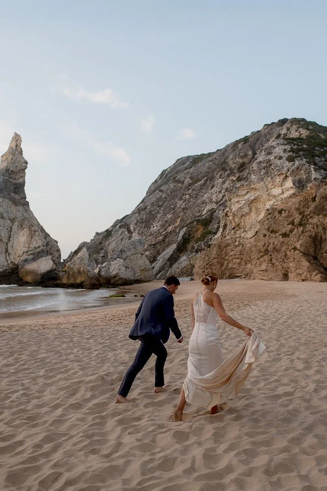 A bride and groom walking barefoot on the beach, with rocky cliffs in the background during sunset or late afternoon.