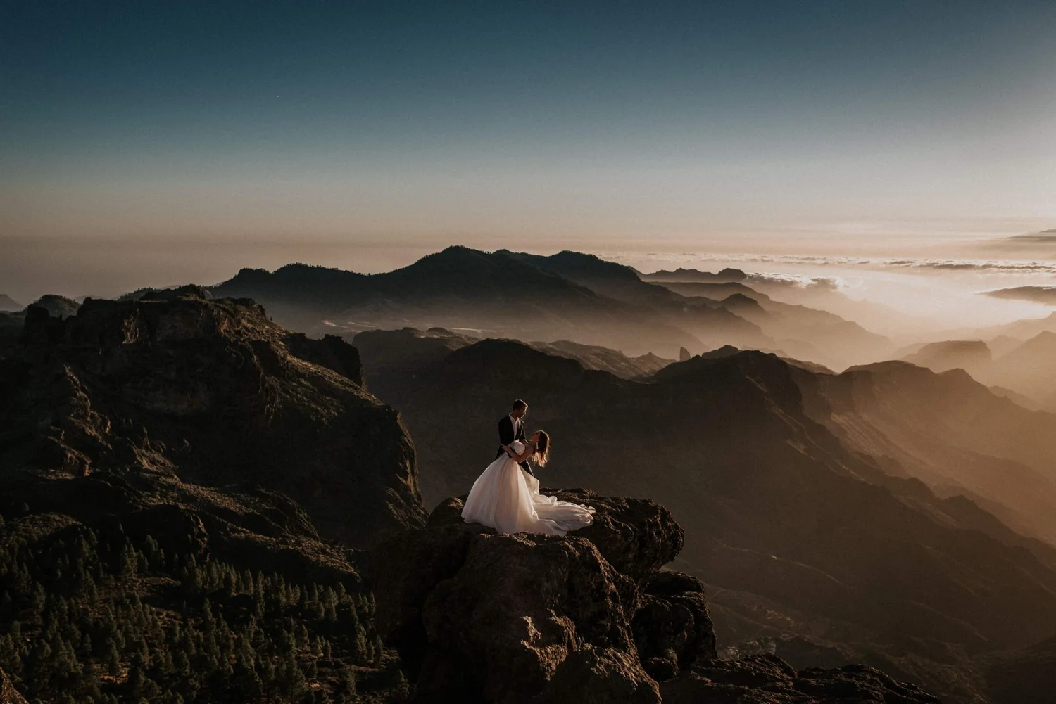 A bride and groom sharing a kiss on a rocky mountain with a landscape of mountains and clouds in the background at sunset.