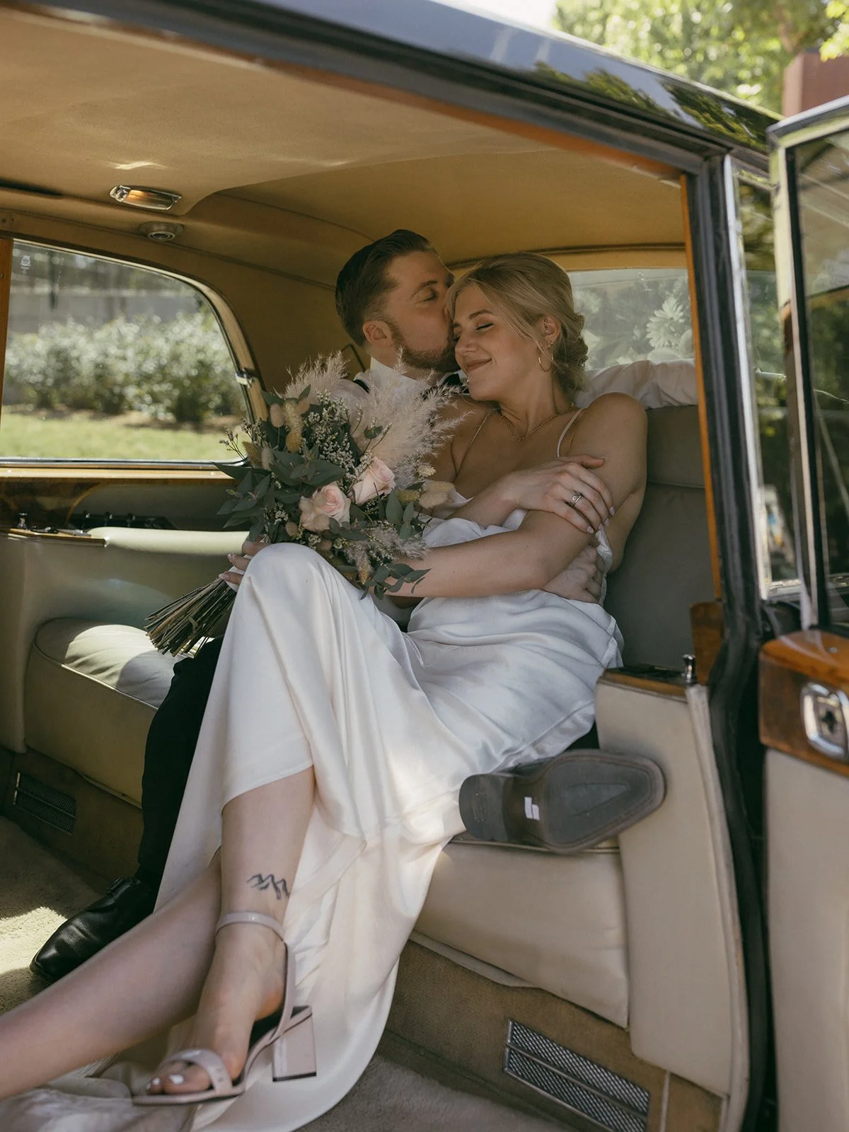 A wedding couple sitting inside a vintage car, sharing a tender moment, with the groom kissing the bride on the forehead. The bride holds a bouquet of flowers and wears a white dress, while the groom wears a suit. Bright outdoor scenery is visible through the car window.