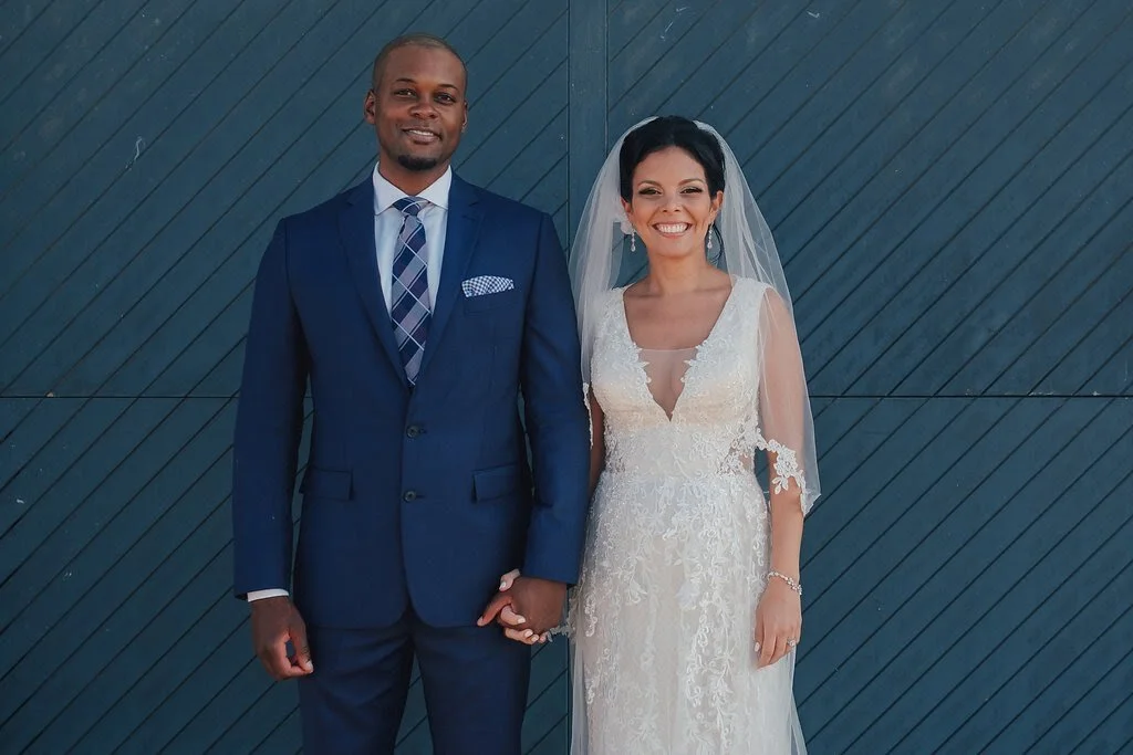 A bride and groom standing side by side, holding hands, posing for a wedding photo in front of a dark blue wooden wall, both smiling.