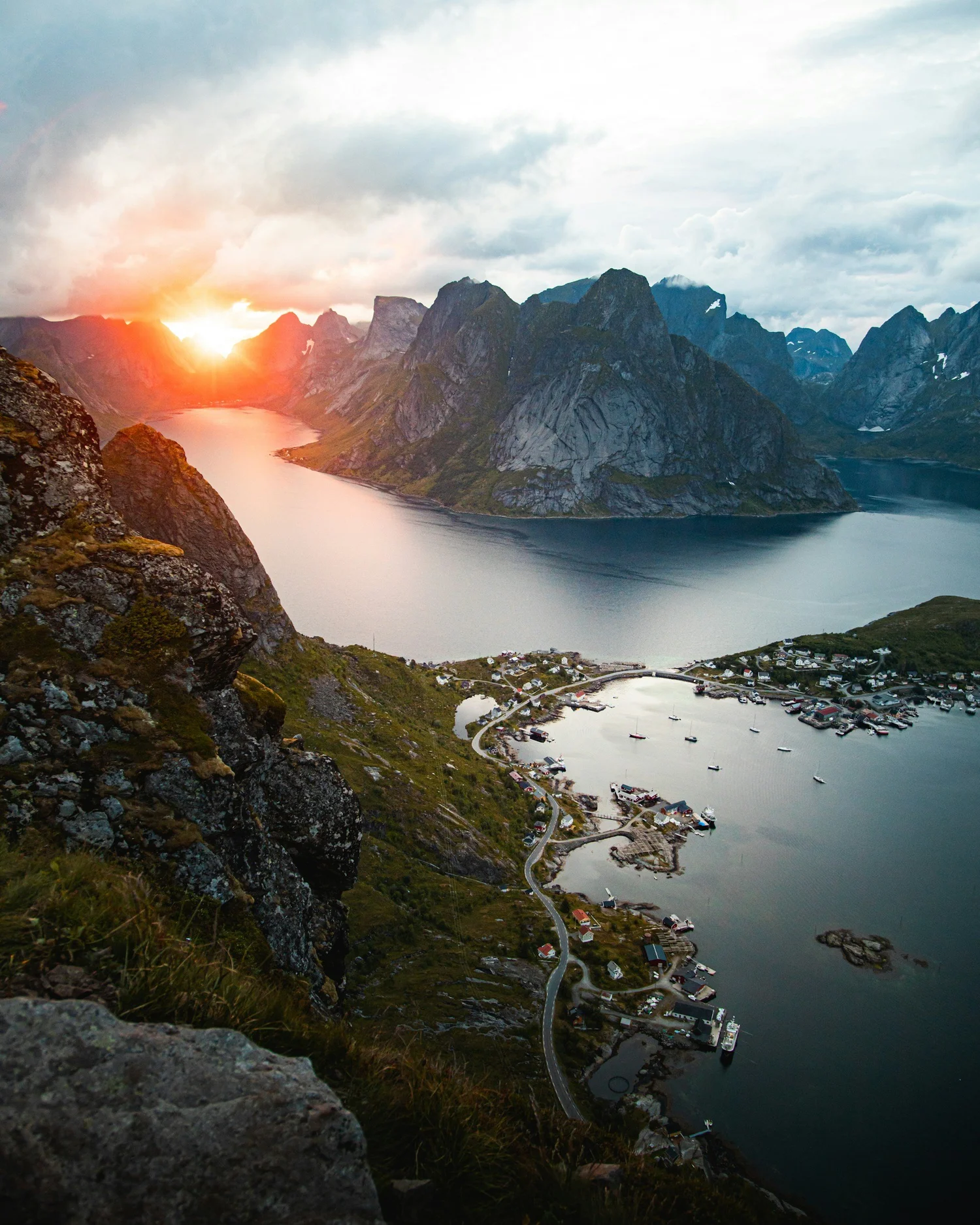 Scenic view of a fjord with rugged mountains, a small village with boats docked, and the sun setting behind the mountains casting a warm glow over the scene.