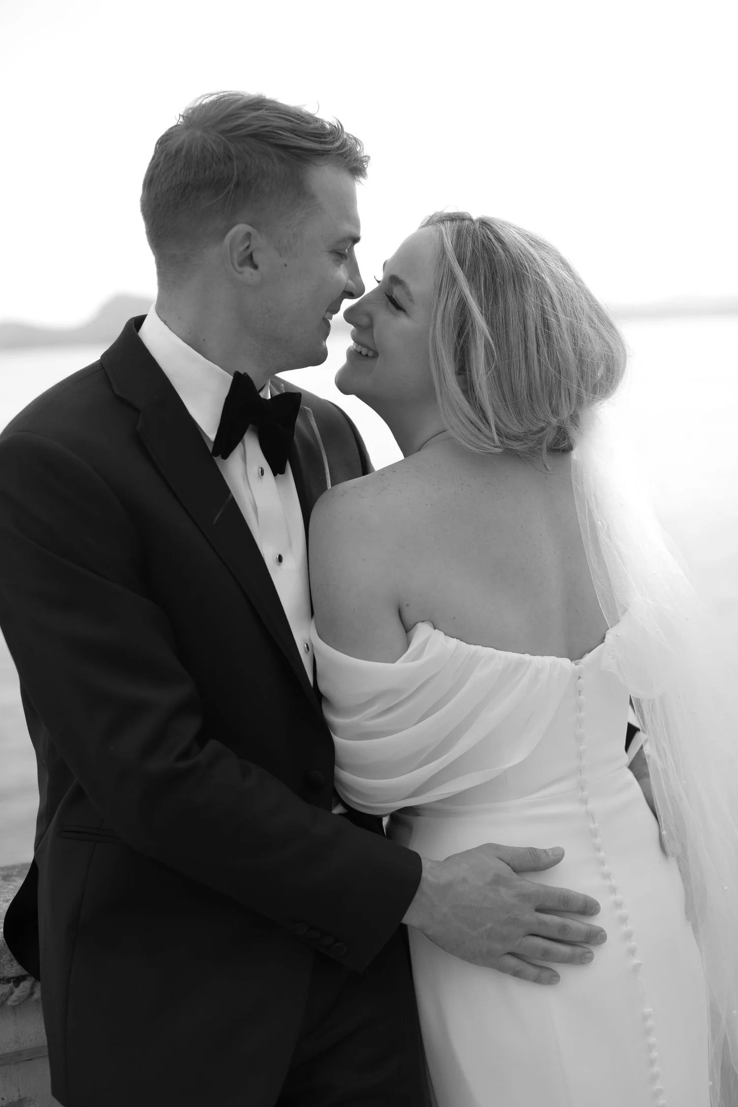 Black and white photo of a happy bride and groom on their wedding day, smiling and touching noses, standing close to each other outdoors.