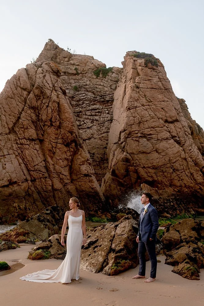 A bride and groom standing barefoot on a beach with large rocks and towering cliffs in the background during a wedding photoshoot.