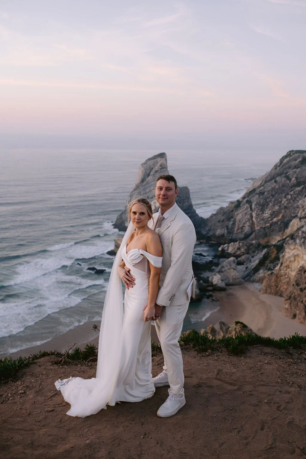 A bride and groom in wedding attire standing on a rocky cliff near the ocean at sunset with large rocks and waves in the background.