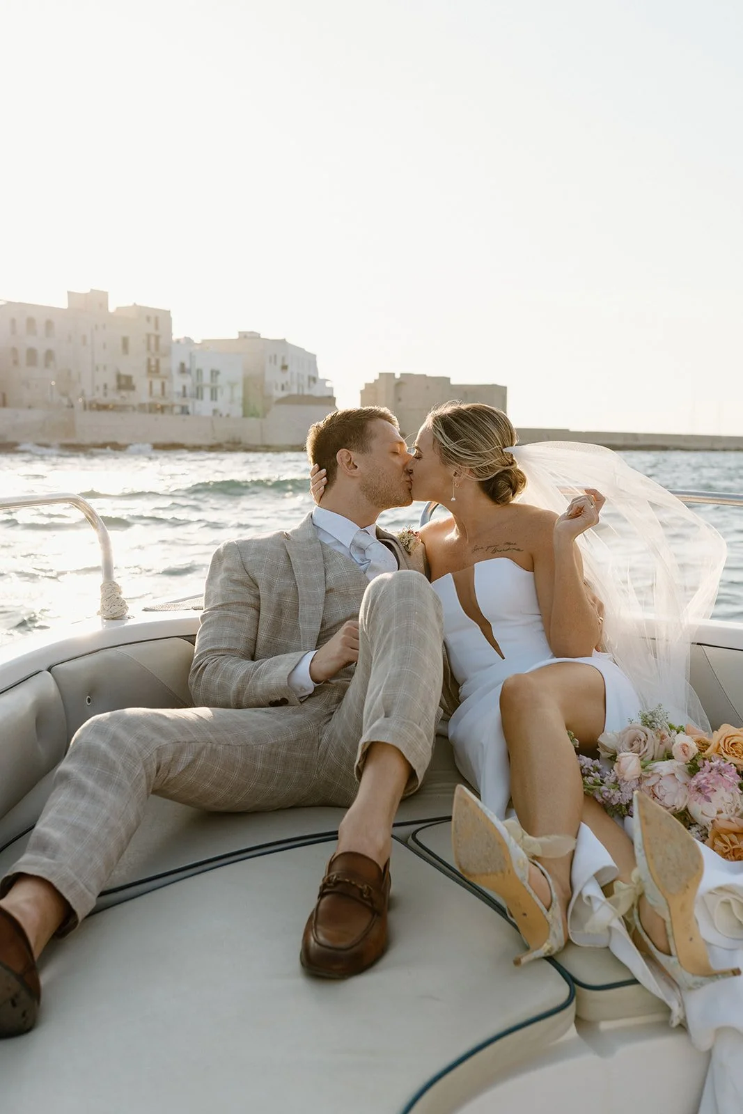 A newlywed couple sharing a kiss on a boat during sunset, with water and buildings in the background.