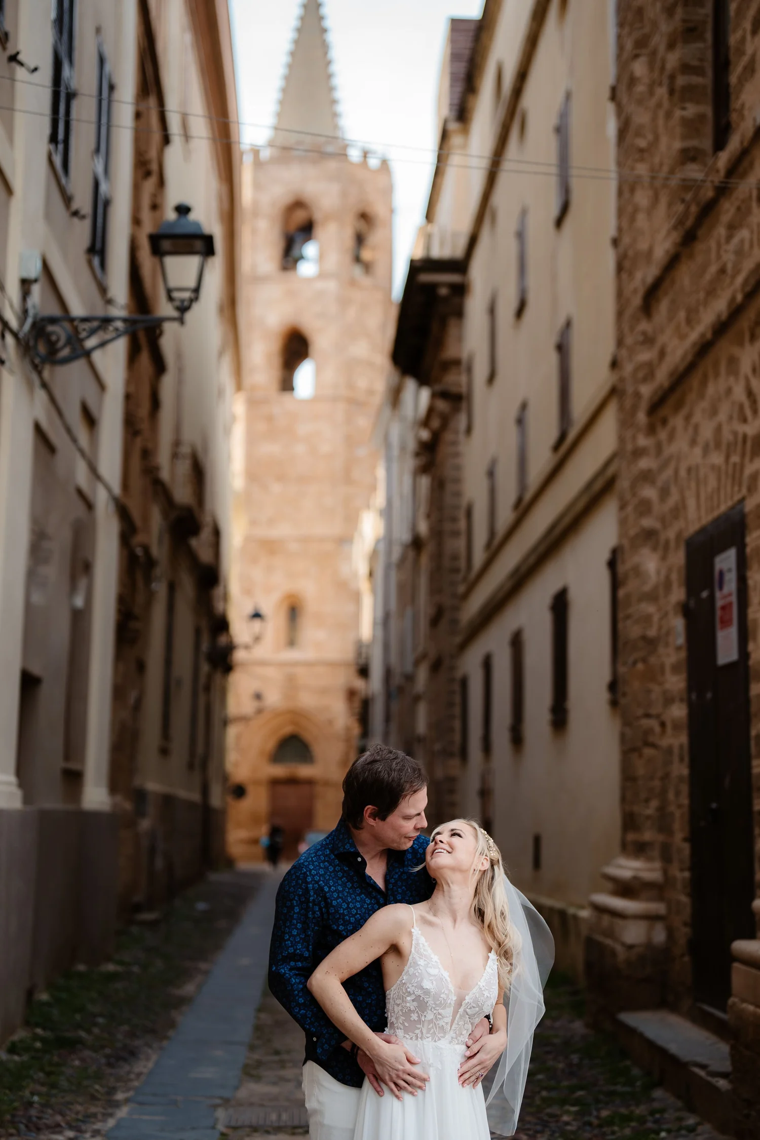 A couple dressed in wedding attire stands close on a narrow historic street with old brick buildings and a church steeple in the background, sharing a tender moment.