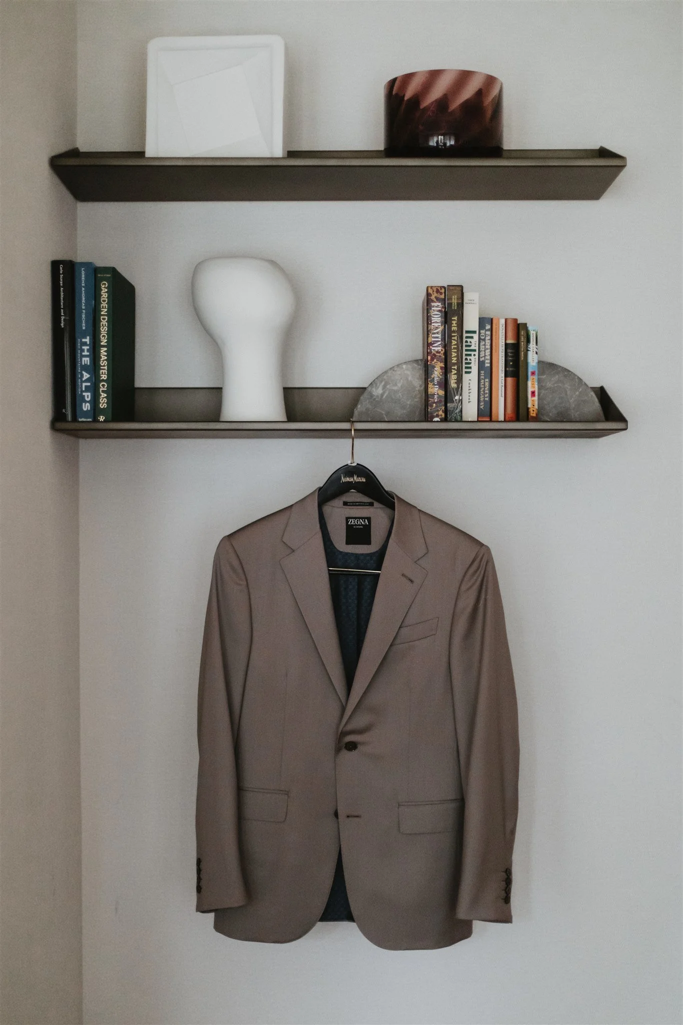 A beige suit jacket hangs on a black hanger on a wall, with two books and a decorative stone placed above it on two shelves.