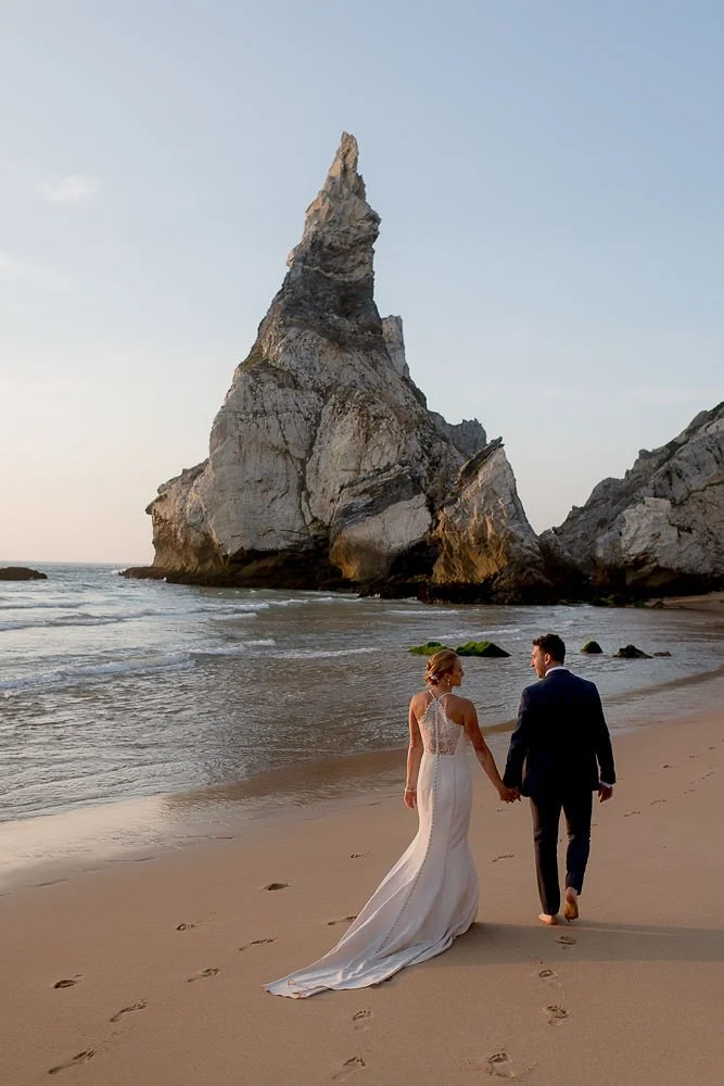 A bride and groom holding hands walking on a sandy beach near a large rock formation during sunset.