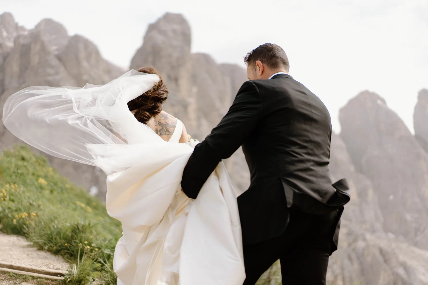 A bride and groom are dancing outdoors with rocky mountains in the background, the bride's veil and dress flowing, and the groom in a black suit.