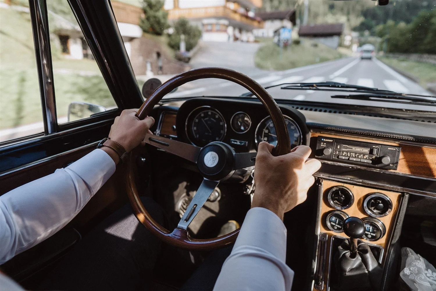 Inside view of a vintage car with a driver's hands on the steering wheel, driving down a rural road with houses and trees in the background.