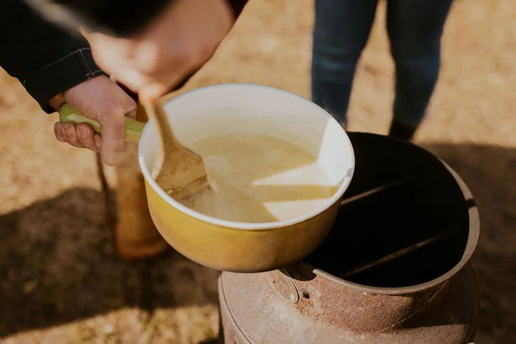 A person stirring a pot of melted cheese with a wooden spatula outdoors.