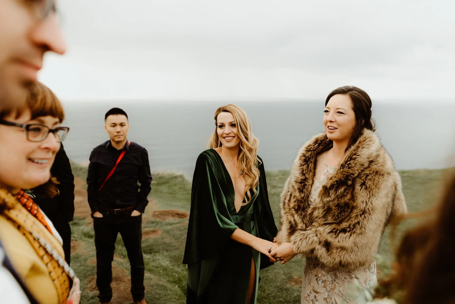 A group of five ethnically diverse people standing outdoors near the coast during daytime, dressed in elegant attire, with one woman wearing a fur coat and another in a dark green gown, engaged in conversation and smiling.
