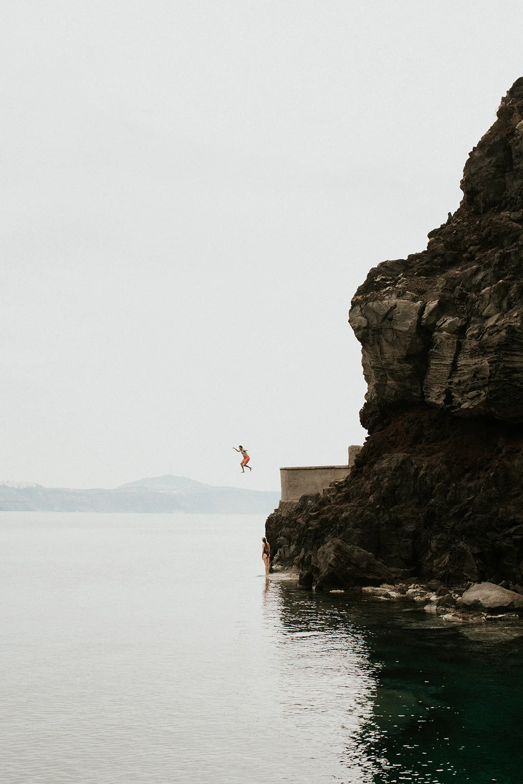 A person wearing orange shorts is jumping from a cliff into water, with another person standing in the water near the cliff, and a large rock formation on the right side of the image, overlooking a body of water with distant mountains in the backgrou