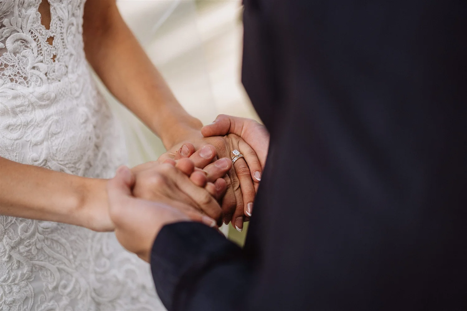 Close-up of a couple holding hands during a wedding ceremony, with the bride wearing a lace dress and the groom in a dark suit, focusing on their hands and wedding rings.