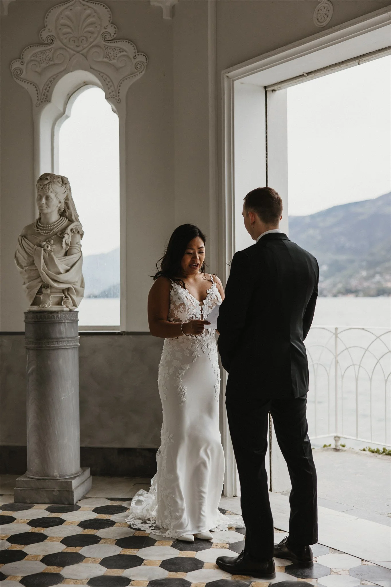 A bride and groom exchanging vows in an elegant, ornate venue with large windows overlooking a mountain landscape. The bride wears a white lace wedding dress, and the groom is dressed in a black suit. A decorative bust sculpture and intricate archite