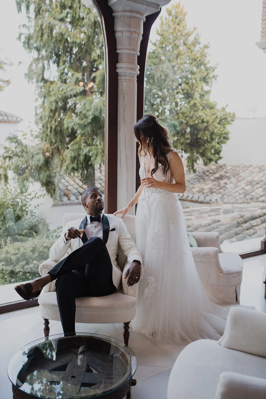 A man in a white tuxedo with a black bowtie sitting on a cream-colored armchair, holding a glass of champagne, looking up at a woman wearing a white wedding dress with lace details, who is standing beside him and also holding a glass of champagne. Th