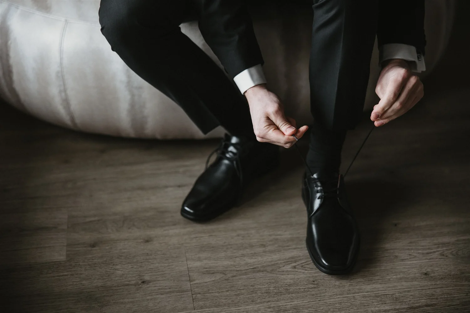 A person dressed in a suit is sitting down, putting on black dress shoes, with a visible polished toe and a long black sock. The person is kneeling on a dark wood floor, near a beige armchair.