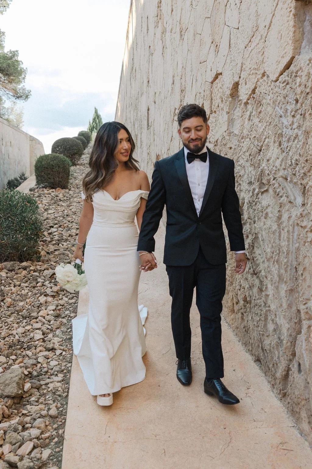 A newlywed couple walking hand in hand outdoors beside a stone wall, with the bride holding a bouquet of white flowers and both dressed in formal wedding attire.