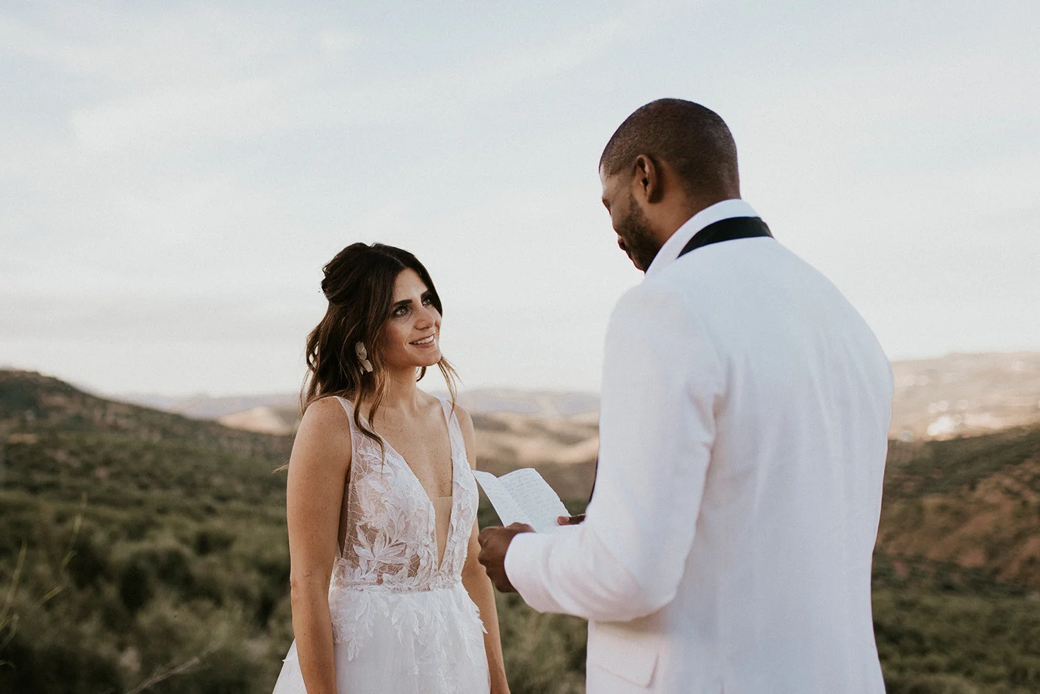 A bride with dark hair in a white lace wedding dress looks at a groom in a white tuxedo during an outdoor wedding ceremony in a scenic hilly landscape.