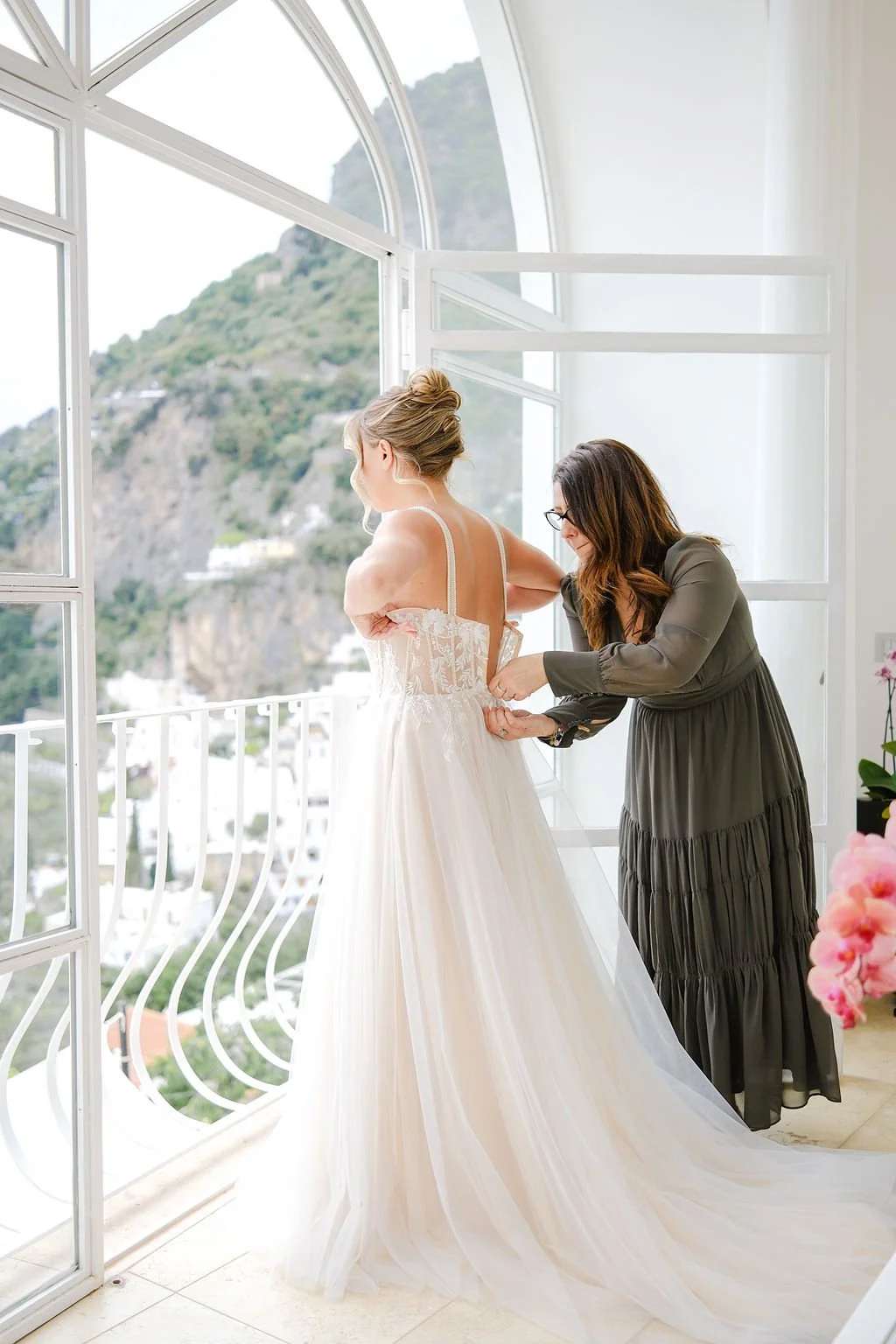 A woman in a wedding dress is being helped with her outfit by another woman in a long gray dress, inside a bright room with large windows overlooking a scenic hillside.