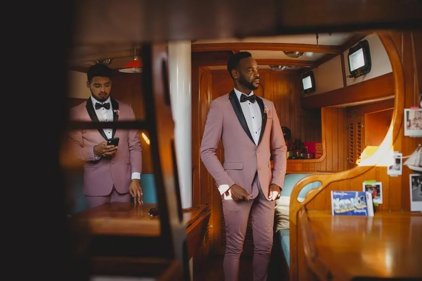 Two men in pink tuxedos with black bow ties in a wood-paneled room, one looking at his phone and the other standing with hands in pockets.