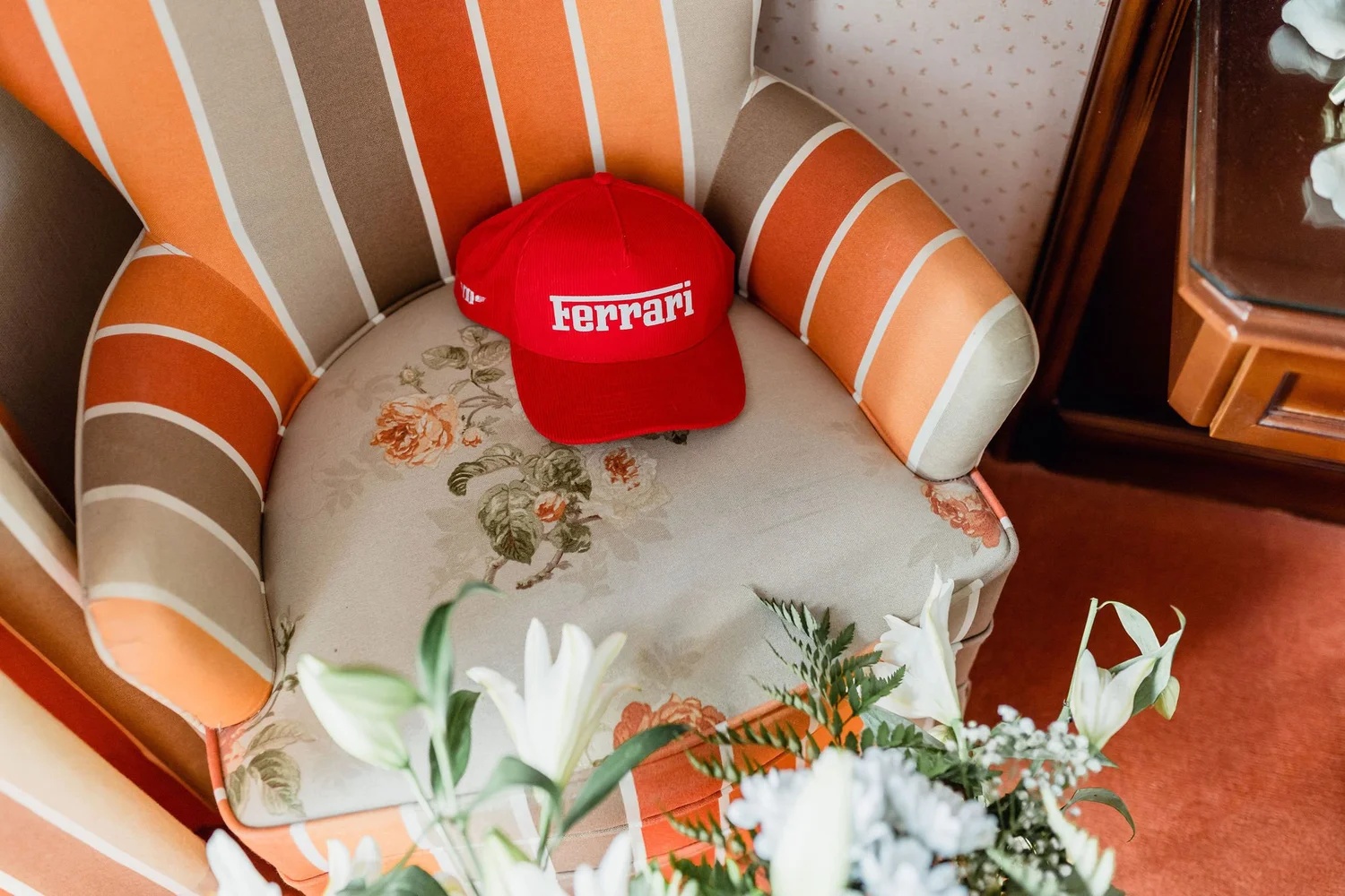 A red Ferrari cap on a vintage floral and striped armchair in a room with patterned wallpaper and a wooden side table with a reflective glass top.