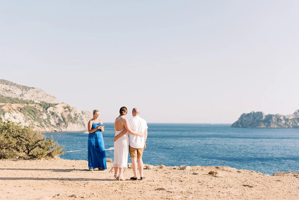 A couple in wedding attire stands on a beach, with a woman in a blue dress nearby, overlooking the ocean and distant cliffs.