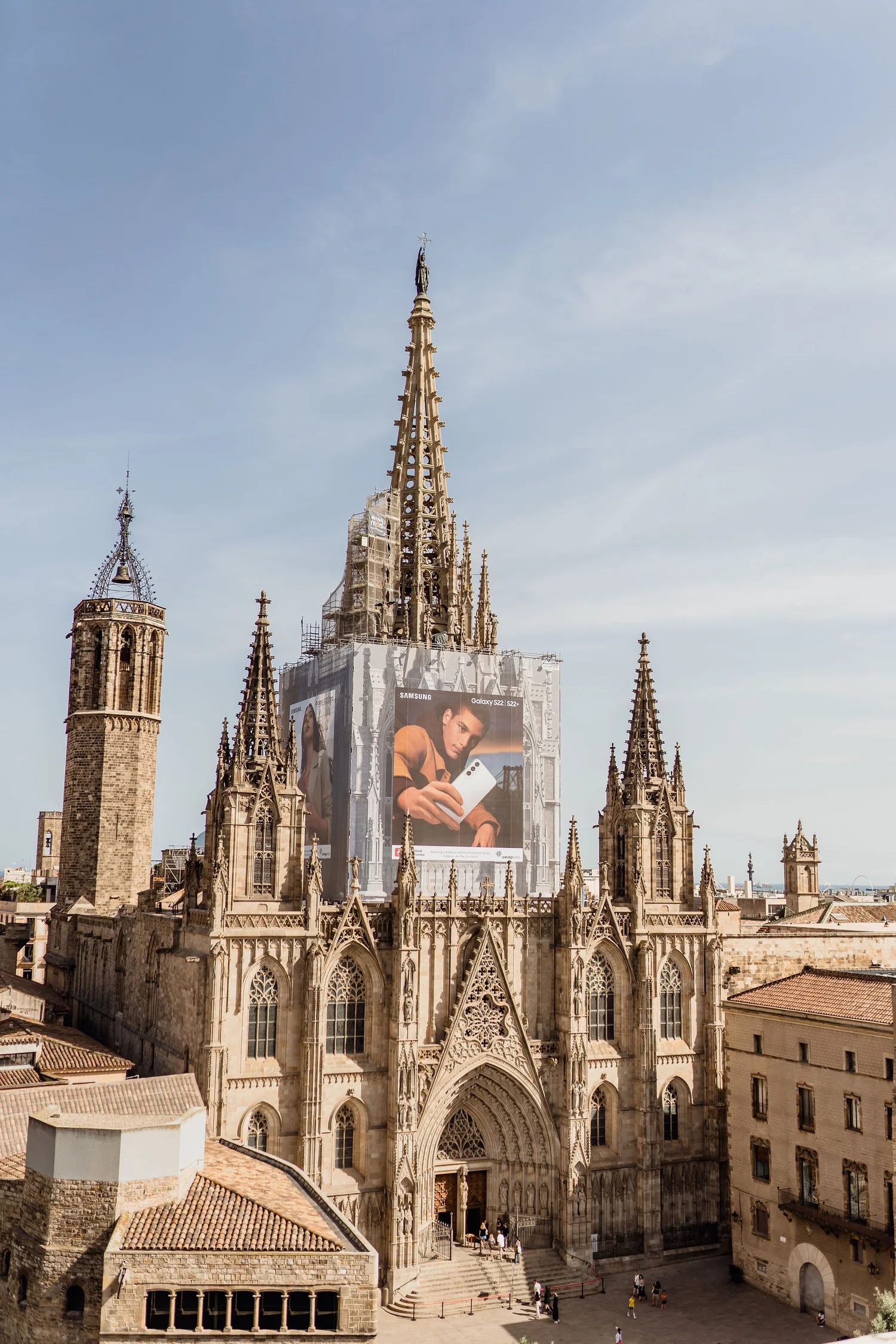A historic Gothic cathedral with intricate stone architecture, large pointed arches, tall spires, and a large digital billboard on the front displaying a fashion advertisement. The sky is clear with some clouds, and people are gathered at the entranc