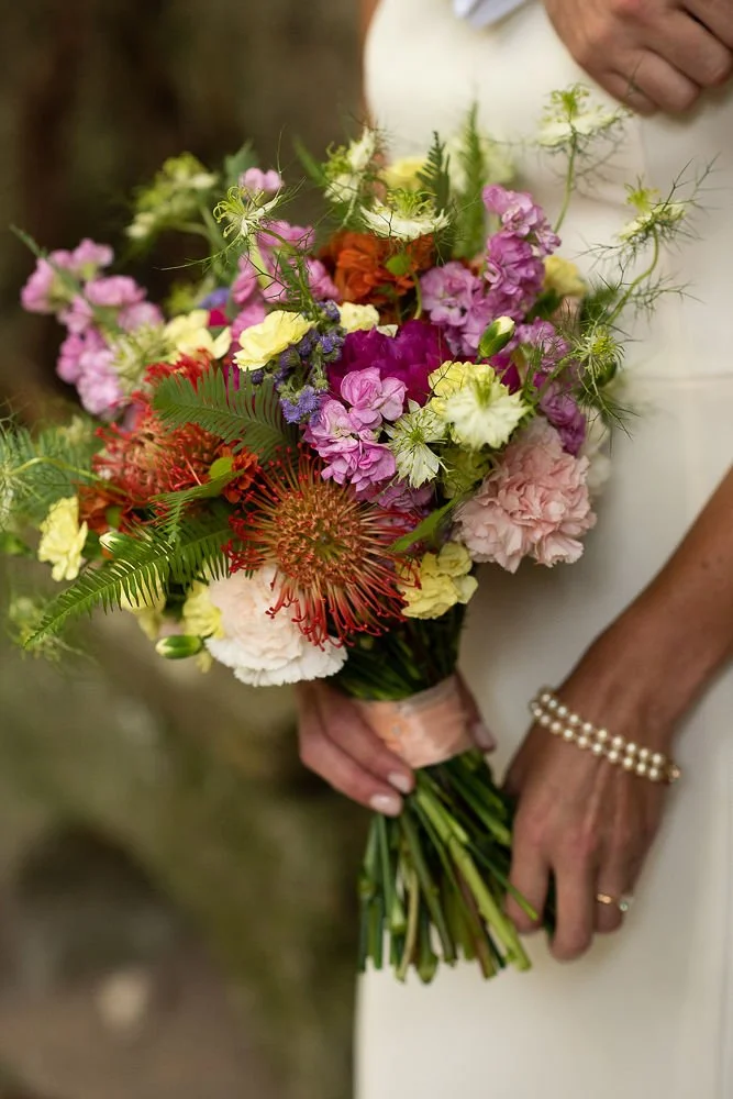A woman holding a colorful bouquet of various flowers, including pink, yellow, orange, purple, and red blooms, with greenery and ferns, wrapped with a peach-colored ribbon.