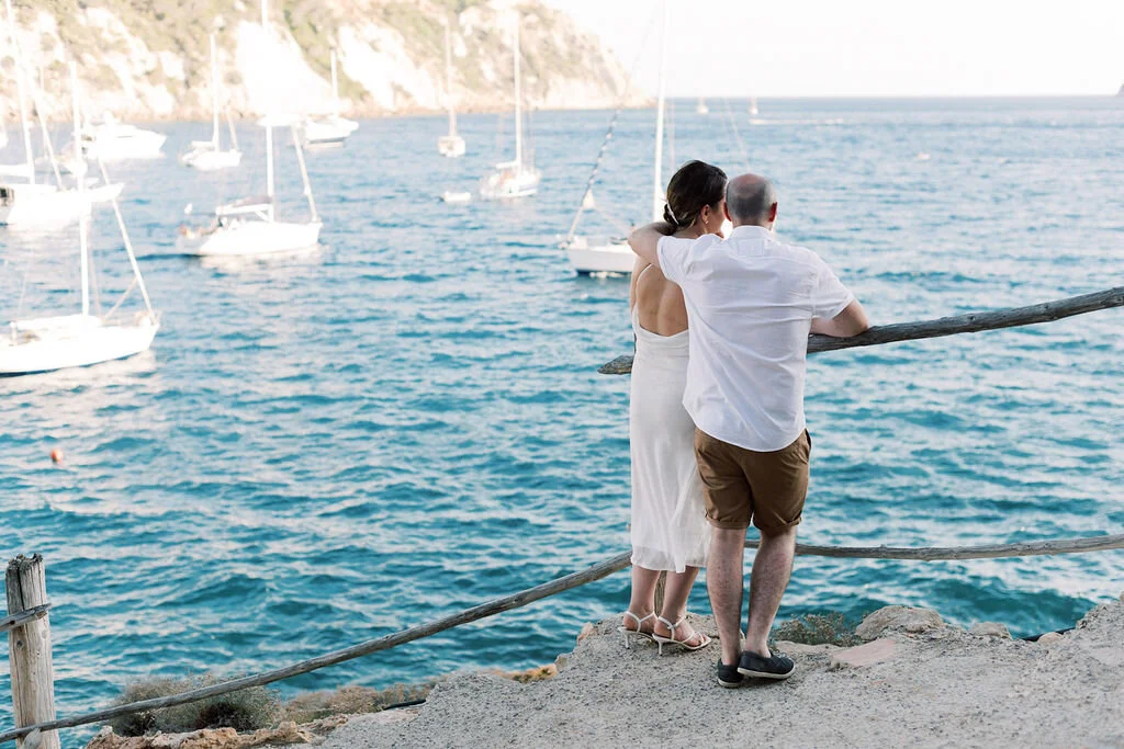 A couple stands on a rocky coast, watching sailboats on the water.