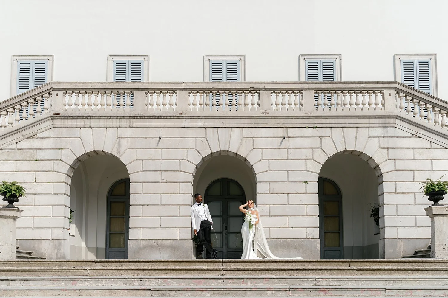 A bride and groom standing on steps in front of a large white building with three arched doorways and four windows with blue shutters.