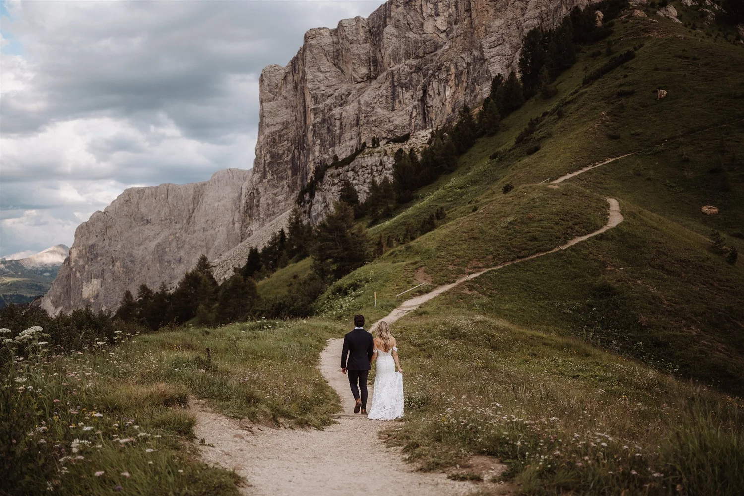 A couple, a man in a suit and a woman in a wedding dress, walking hand in hand along a dirt path through a green meadow surrounded by mountains, with a cloudy sky overhead.