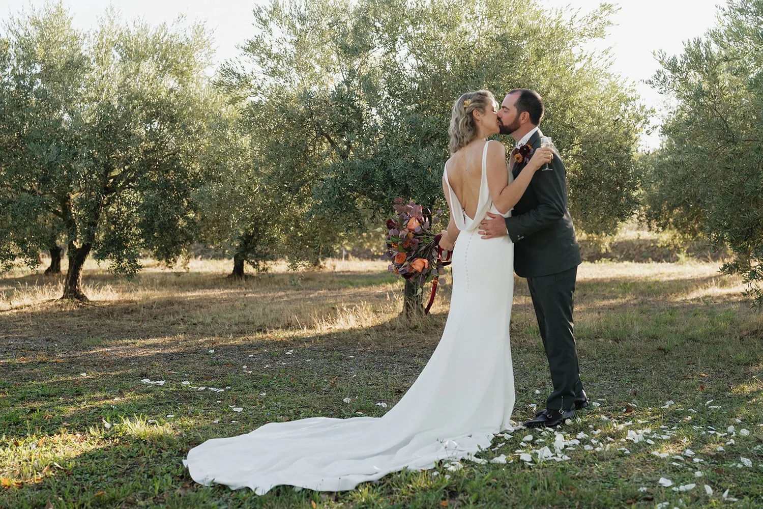 A bride and groom kissing outdoors in a grassy area with trees and sunlight, the bride in a white gown holding a bouquet, the groom in a black suit.
