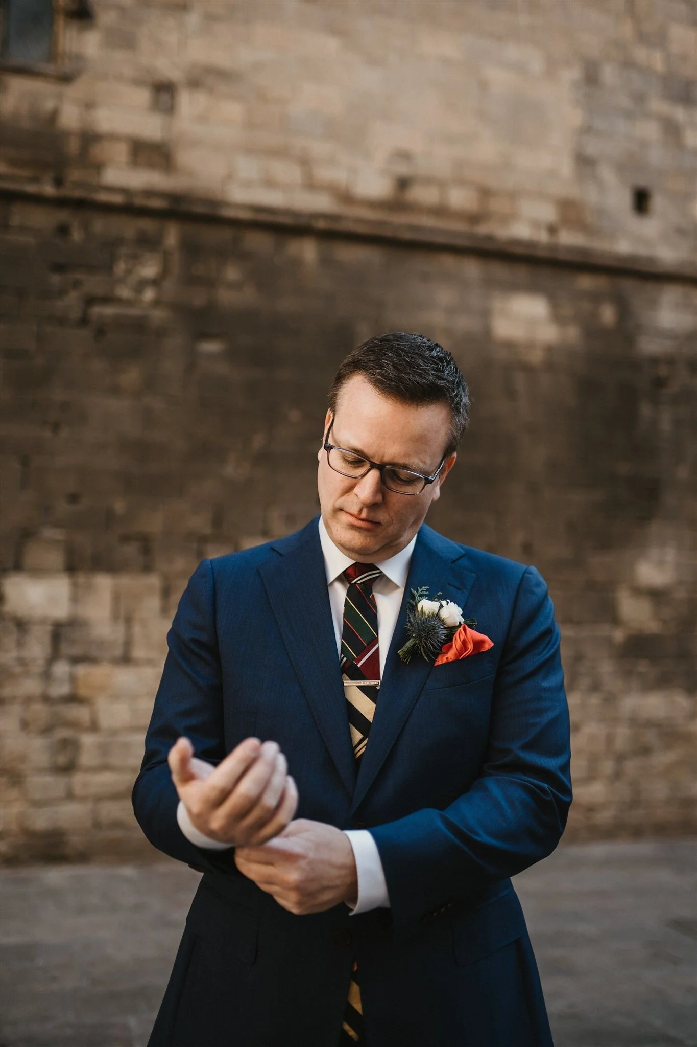 A man wearing a blue suit, glasses, and a striped tie, standing against a brick wall background, looking down and adjusting his sleeve.