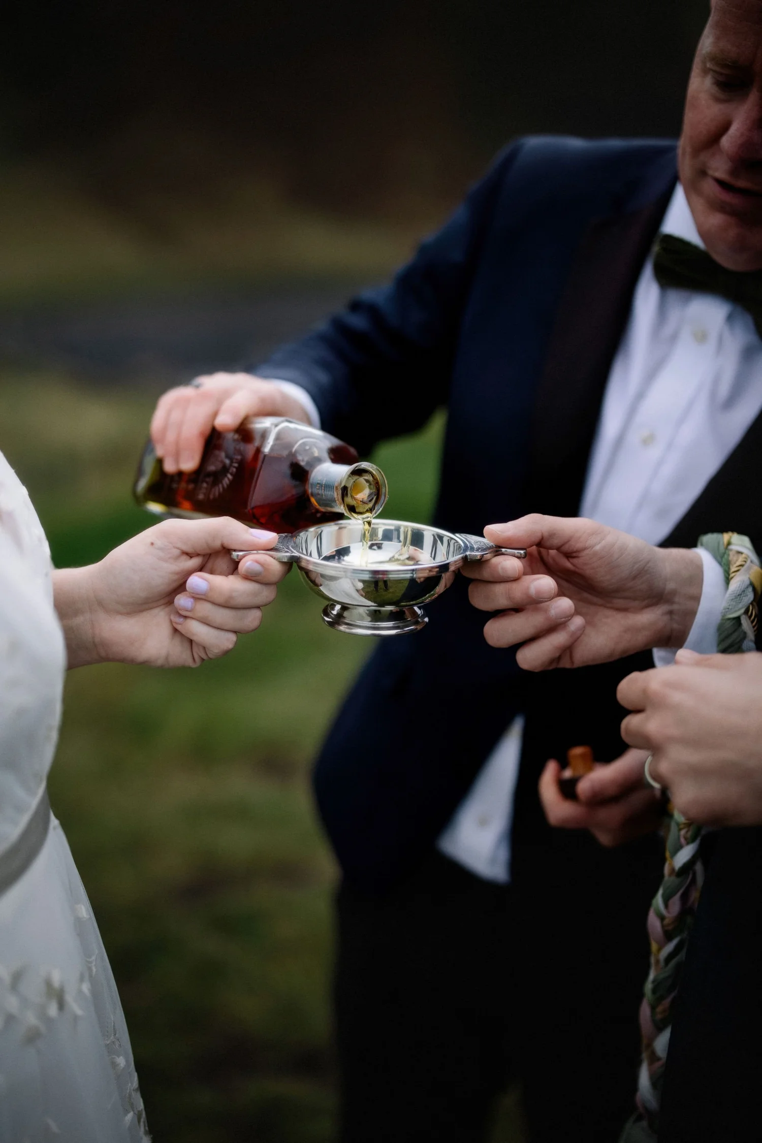 A person in a dark suit and bow tie is pouring a brown liquid from a bottle into a silver tray held by another person in a white dress.