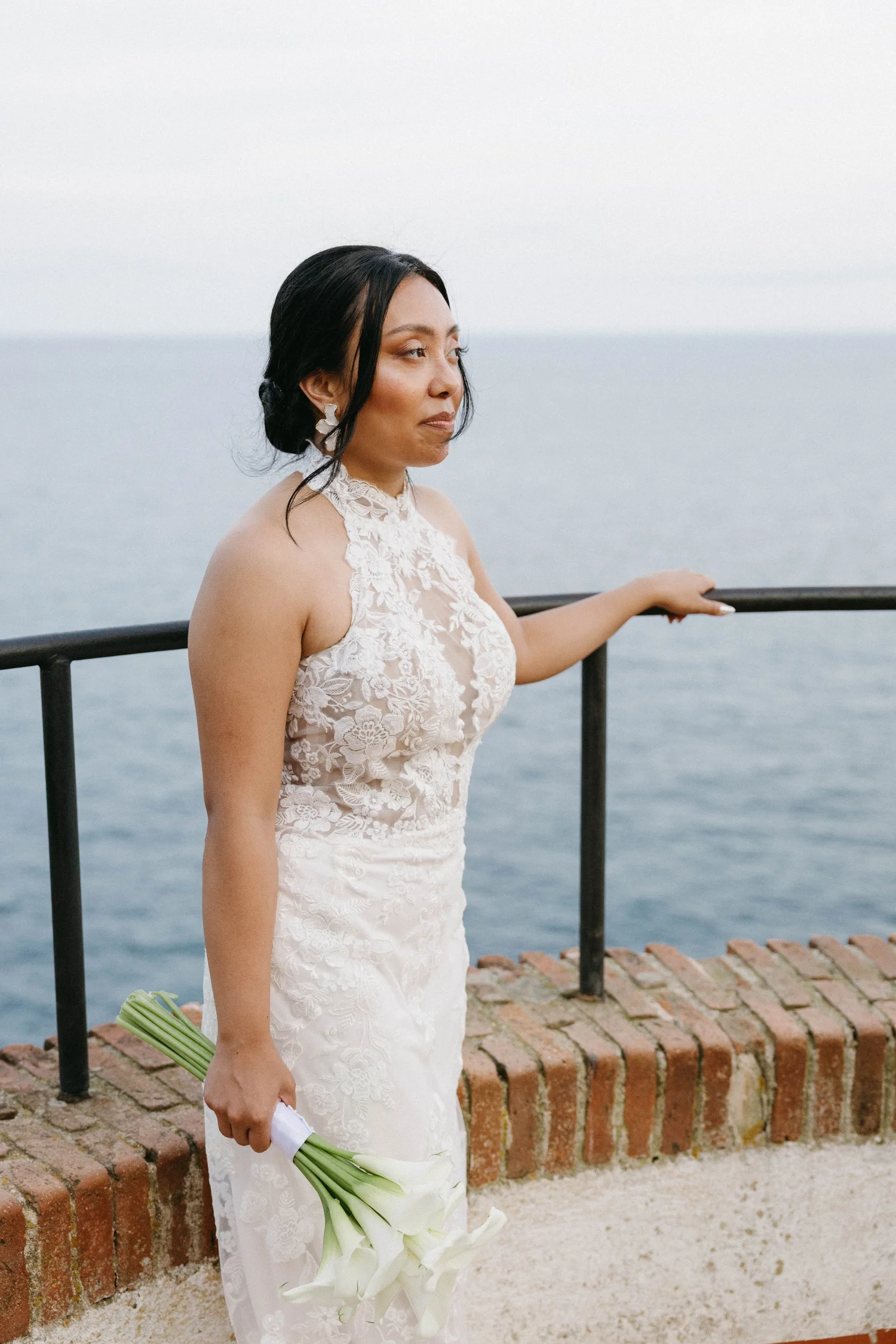 A woman in a wedding dress holding a bouquet of white calla lilies, standing by a brick wall near the water with her hand resting on a black metal railing.