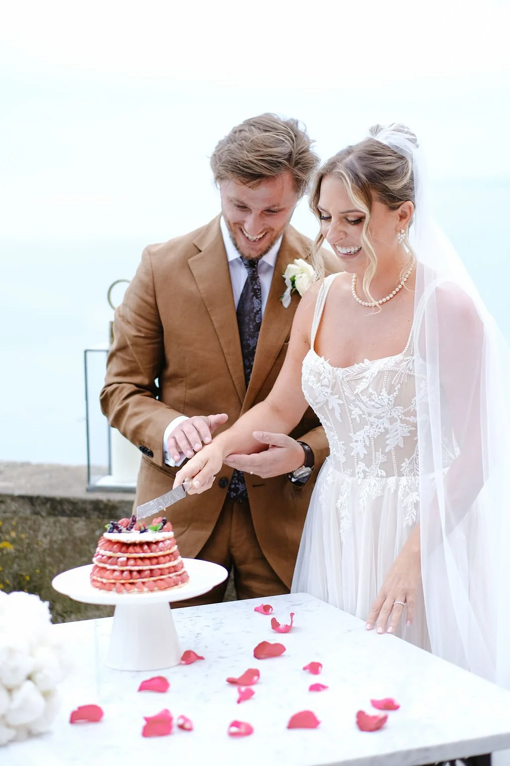 A bride and groom smiling as they cut a wedding cake outdoors, surrounded by rose petals.