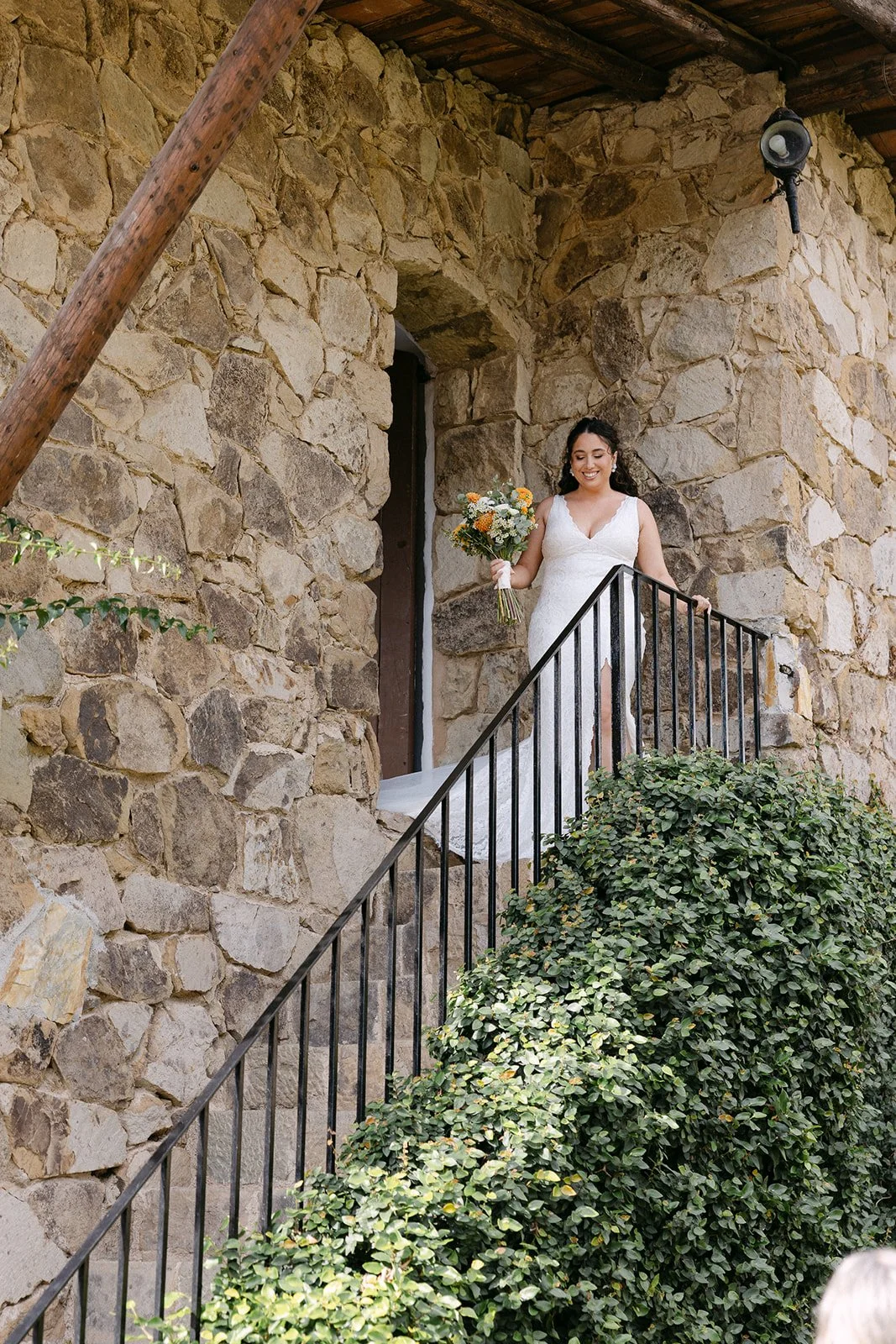 A bride in a white lace wedding dress holding a bouquet of flowers stands on a small staircase outside a stone building, smiling.