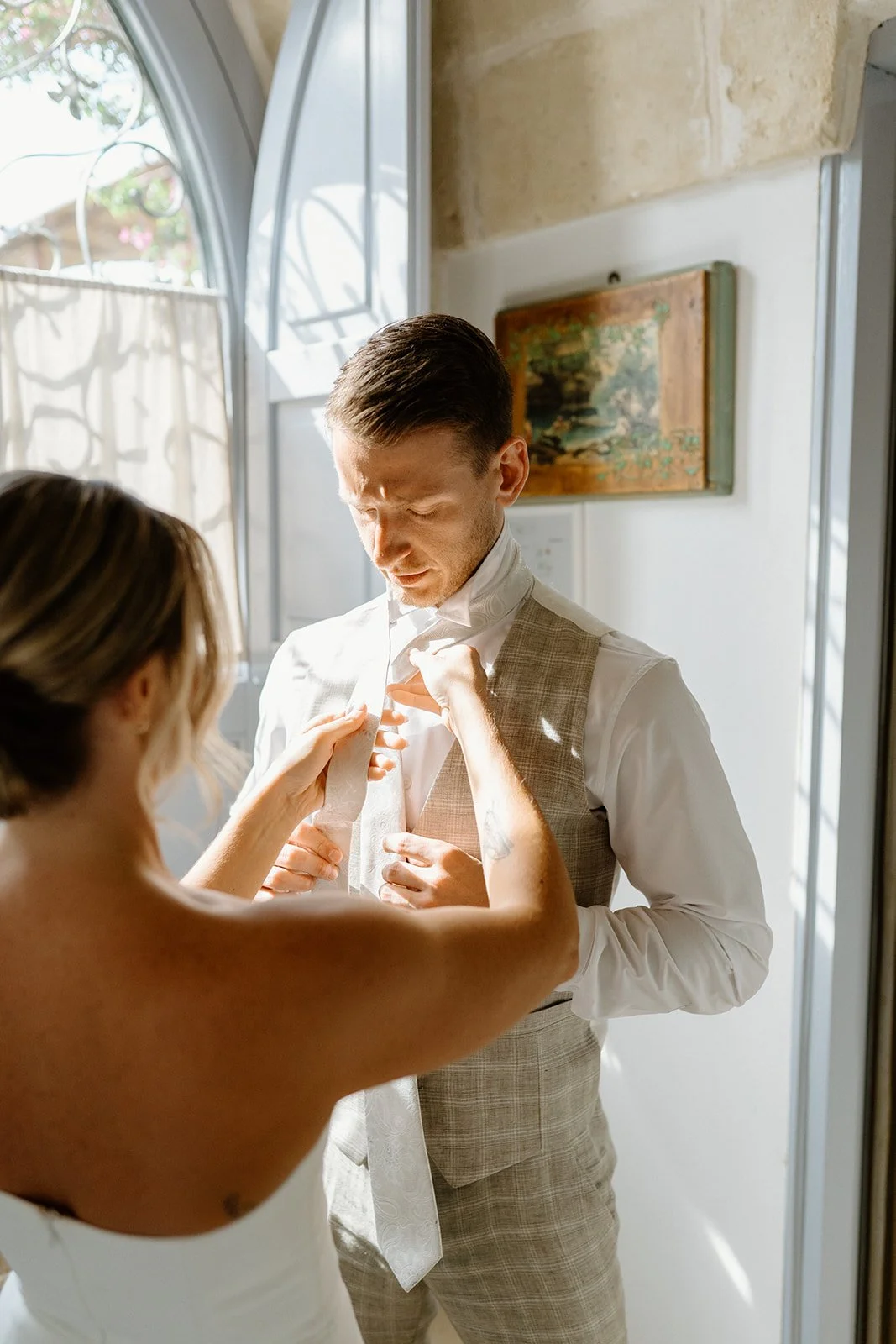 A woman adjusts a man's tie as he prepares for a formal event, in a sunlit room with vintage decor.