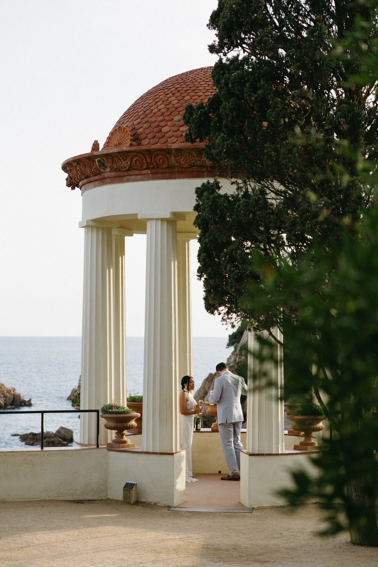 A couple getting married outdoors on a terrace overlooking the ocean, under a cream-colored pergola with red tile roof, columns, and decorative moldings, surrounded by potted plants and trees.