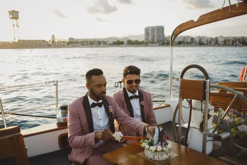 Two men in pink tuxedos cutting a wedding cake on a boat during sunset with a city skyline in the background.