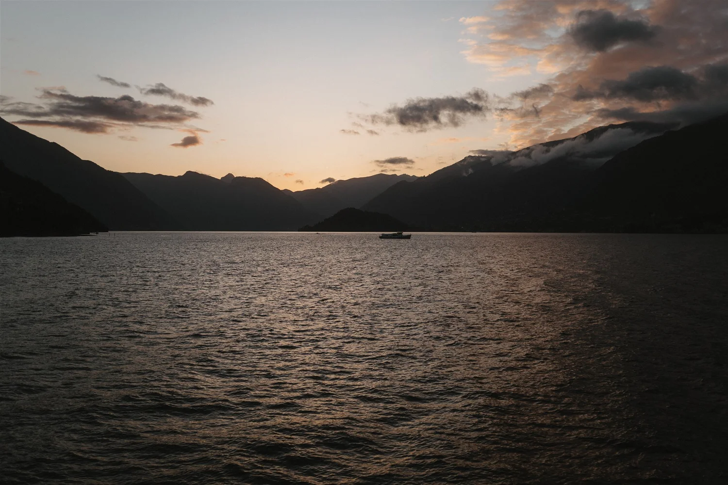 A scenic view of a lake surrounded by mountains at sunset with clouds in the sky.