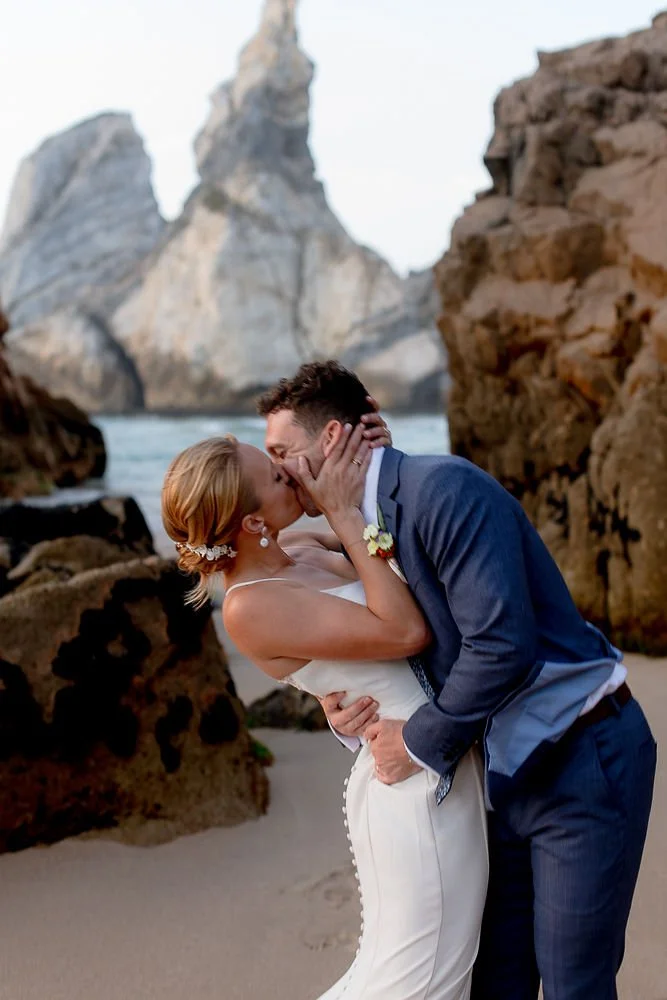 A couple in wedding attire shares a kiss on a beach with large rocks and cliffs in the background.