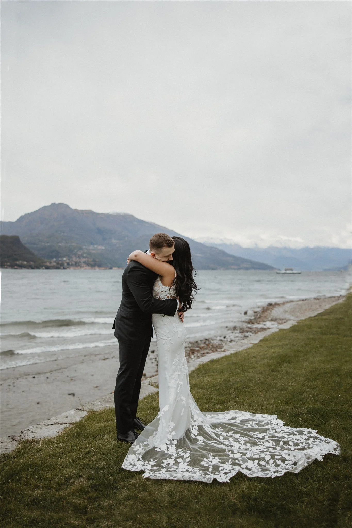 A bride and groom embrace on a grass and beach shoreline with mountains in the background, cloudy sky, and a boat on the water.