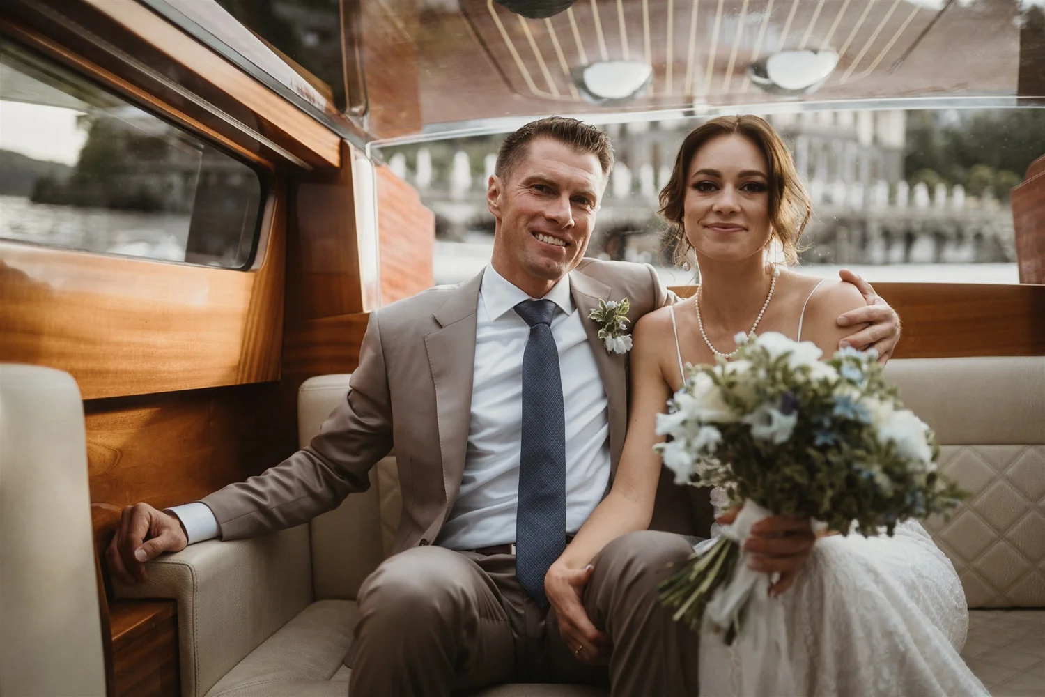 A newlywed couple sitting in a boat, smiling, with the bride holding a bouquet of flowers, in front of a riverside scene.