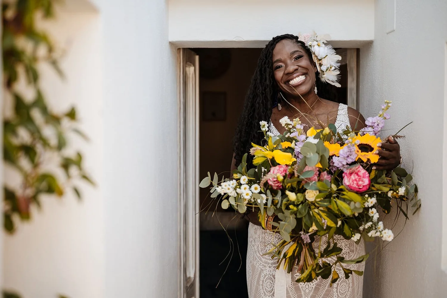 A woman with dark skin and long curly hair stands in a doorway holding a large bouquet of colorful flowers, smiling widely. She is wearing a white lace dress and a floral hair accessory.