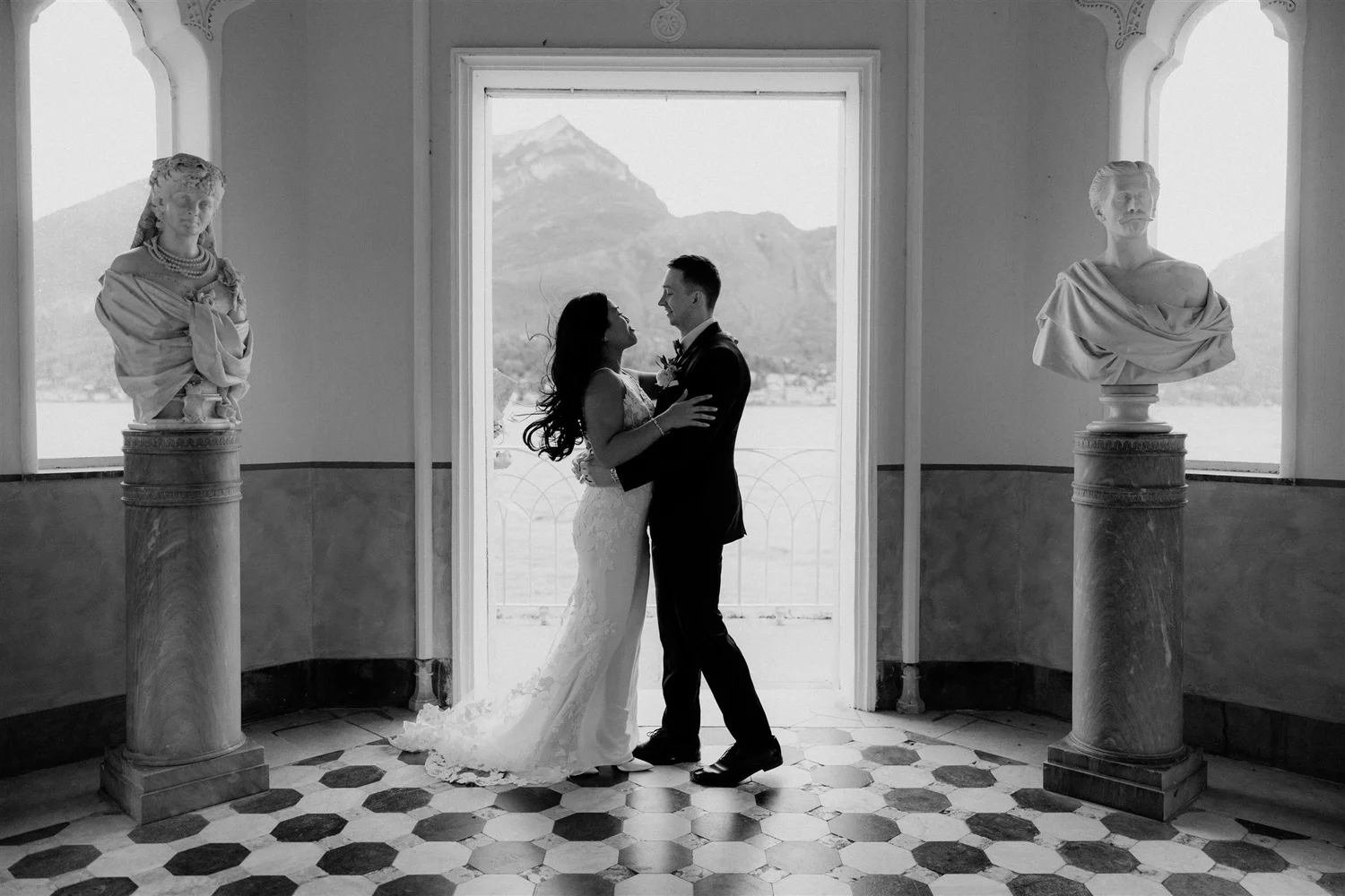 A black-and-white photo of a bride and groom standing close together, indoors by a large window with mountain scenery in the background. The bride is wearing a wedding dress with lace details, and the groom is in a suit. Two classical statues on pede
