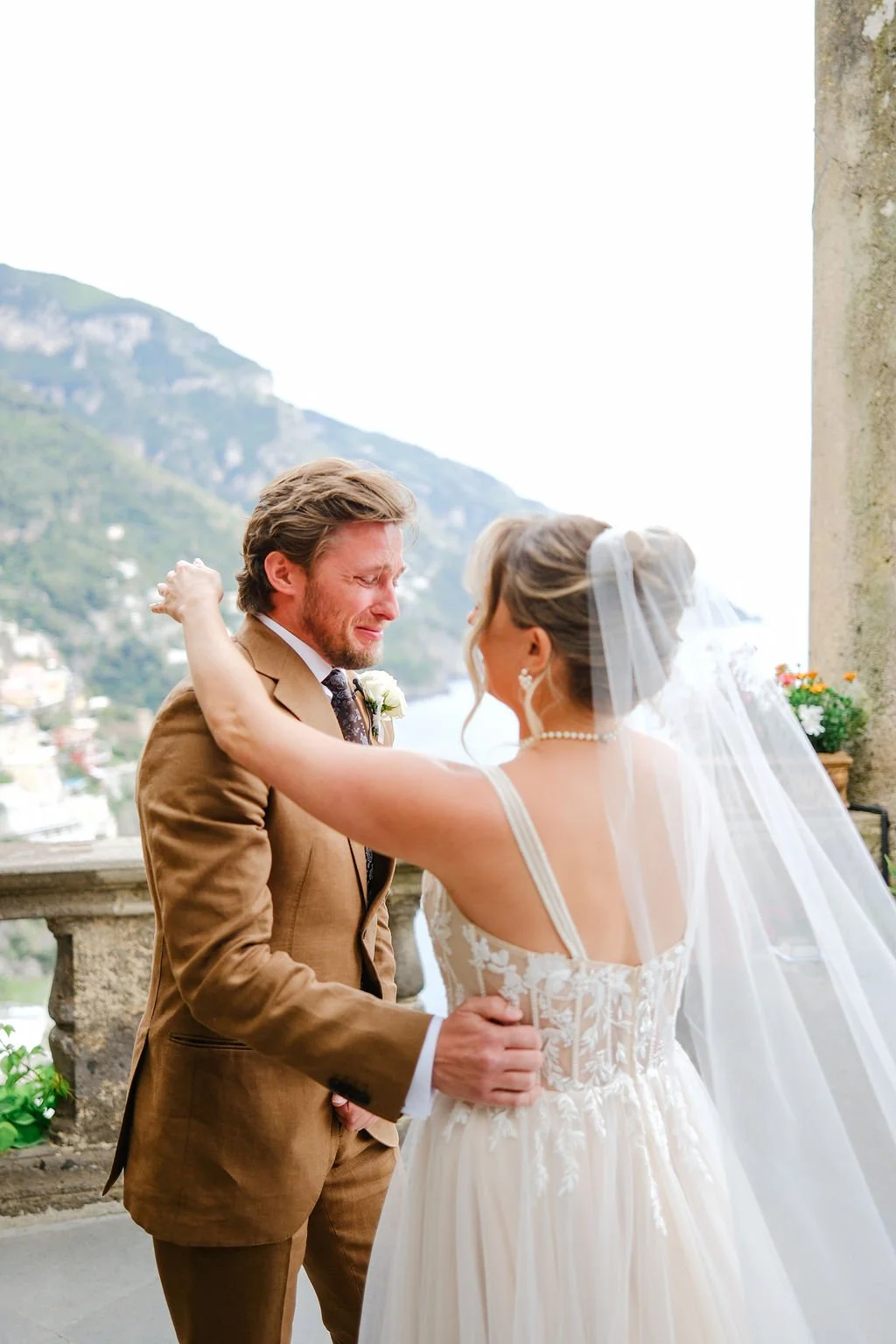 A bride and groom sharing an emotional moment during their wedding ceremony outdoors, with mountains and a coastal town in the background.